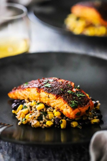 A slice of a blackened salmon fillet sitting on a corn and black bean salad on a black dinner plate.