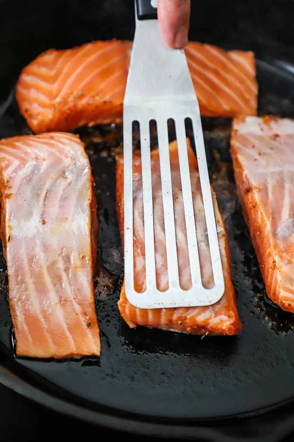 A thin metal spatula pressing down on a salmon fillet that is being seared in cast-iron skillet along with three other fillets.