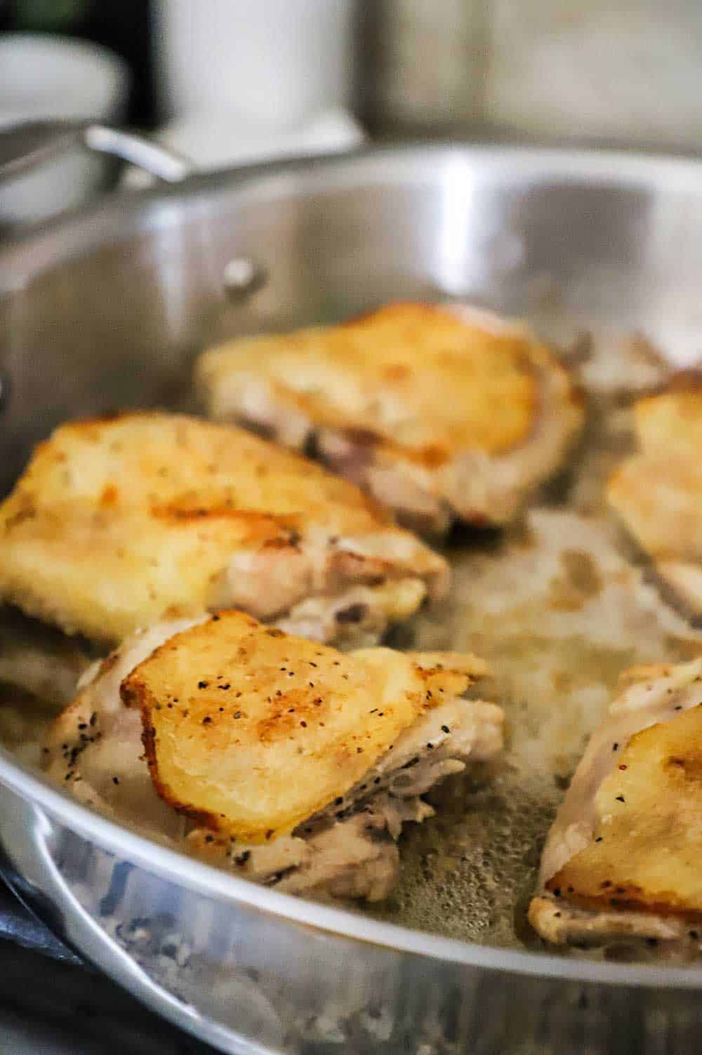 A large stainless steel skillet filled with skin-on chicken thighs that have been seared causing the skin on top to be crispy.