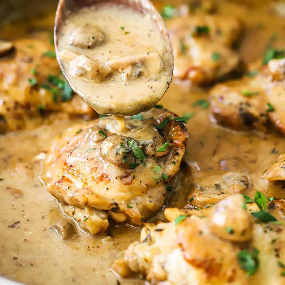 A wooden spoon being used to pour a creamy mushroom gravy sauce over seared chicken thighs.