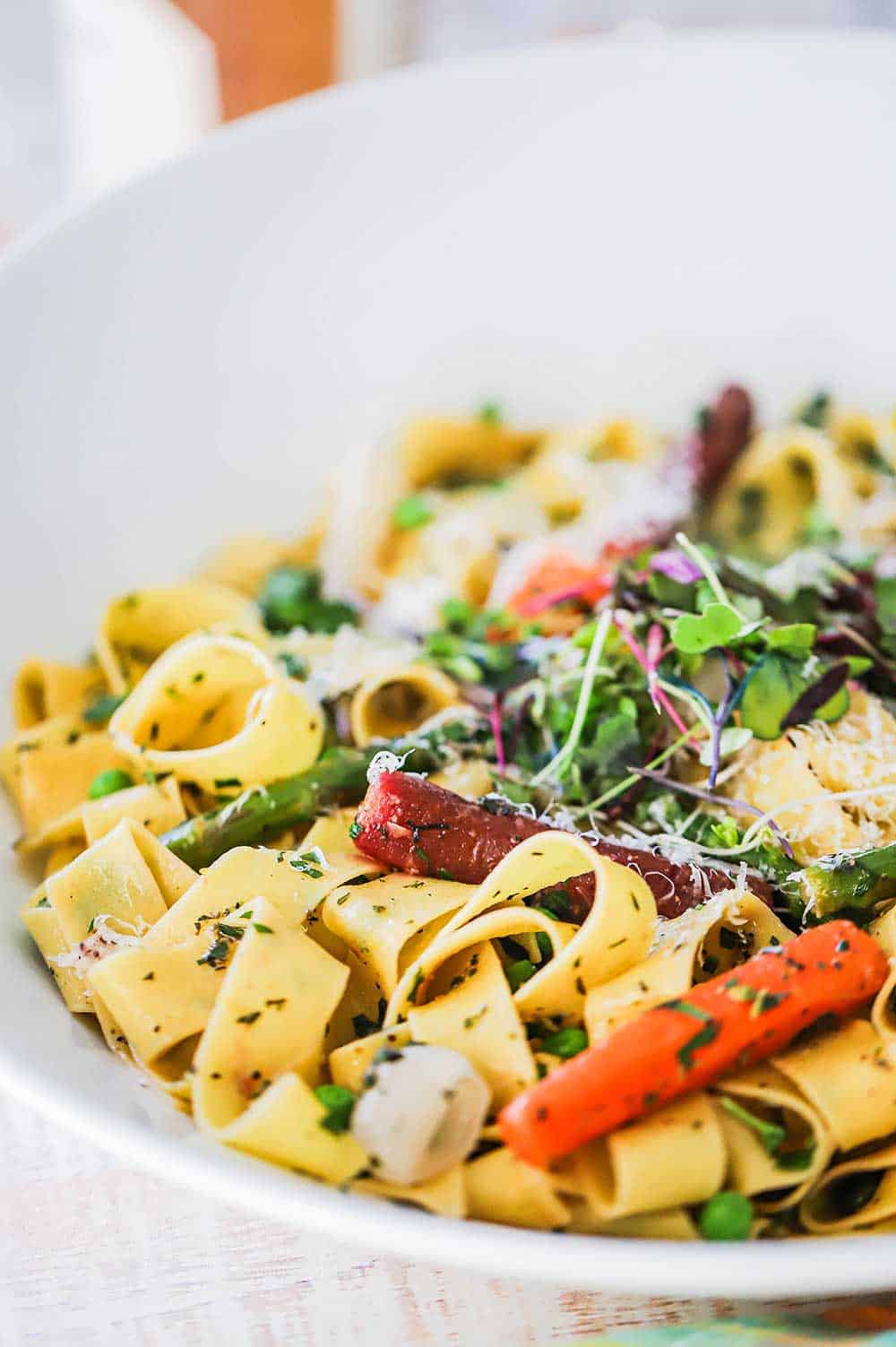 A large white serving bowl filled with fresh pappardelle pasta and sautéd spring vegetables.