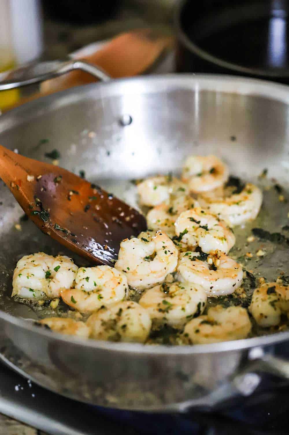 A wooden spoon stirring shrimp that has been sautéd with herbs in a large stainless steel skillet. 