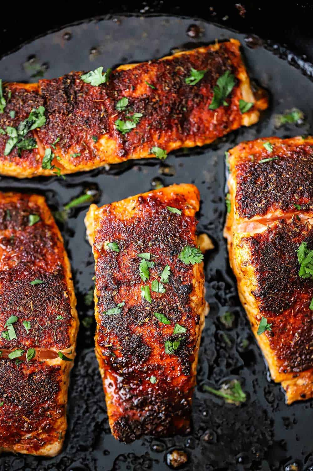 Four blackened salmon fillets sitting in a sizzling black cast-iron skillet all garnished with fresh chopped parsley.