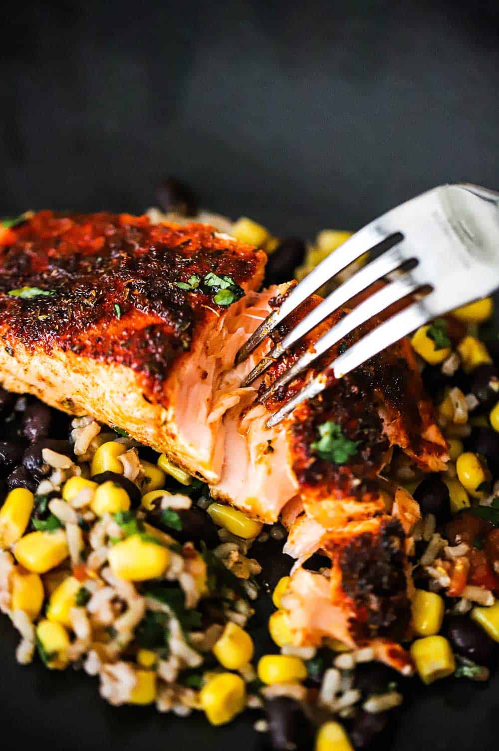 A fork flaking open a fillet of salmon that is blackened and sitting on a corn and black bean salad on a black dinner plate.