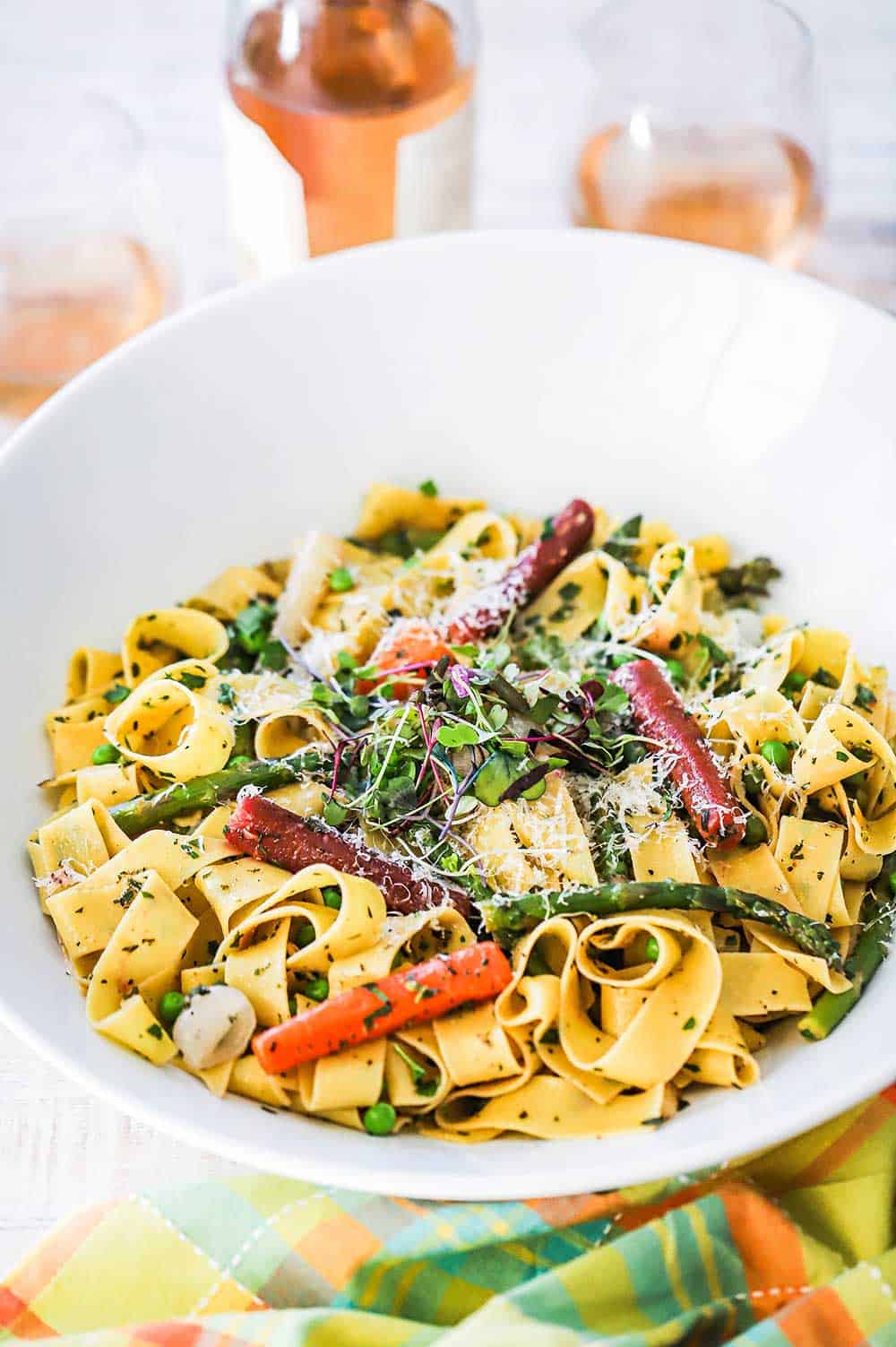 A circular white serving bowl filled with pappardelle with seasonal vegetables next to a bottle of Rosé and two glasses of wine.