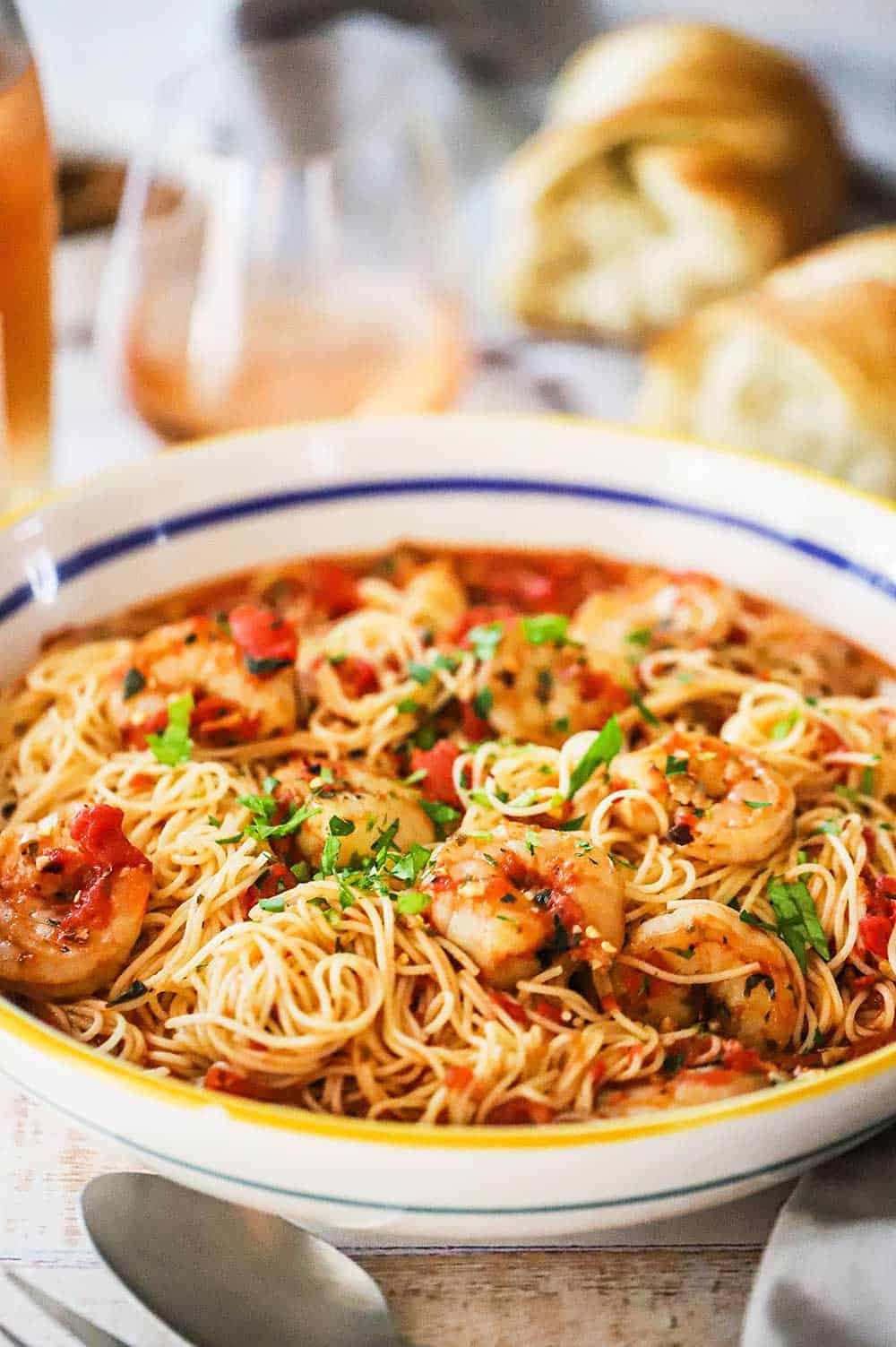 A large pasta bowl filled with shrimp marinara next to a glass of rosé wine and a torn loaf of bread. 