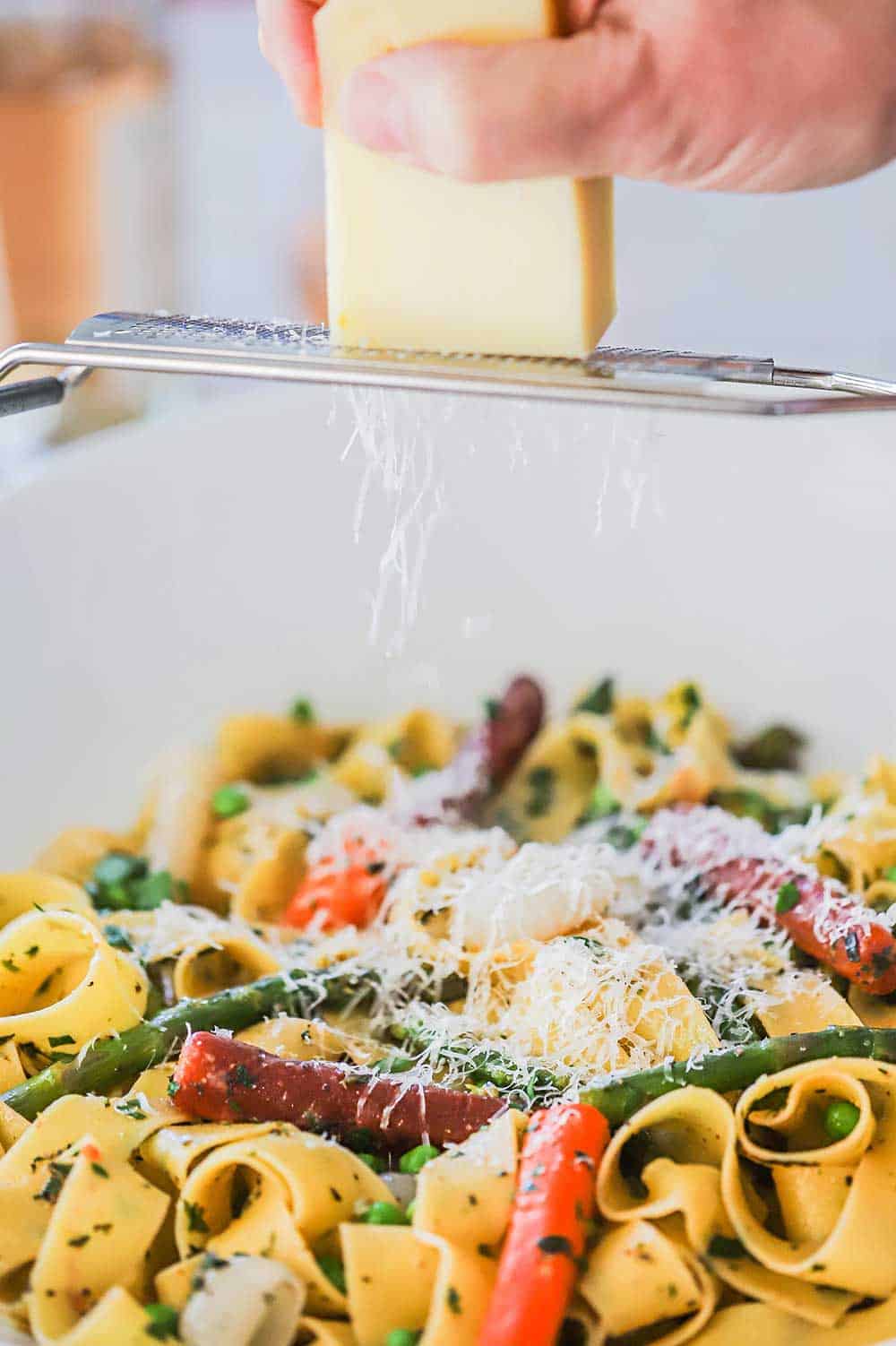 A person grating a block of Parmesan cheese over a bowl filled with fresh pasta and sautéd vegetables.