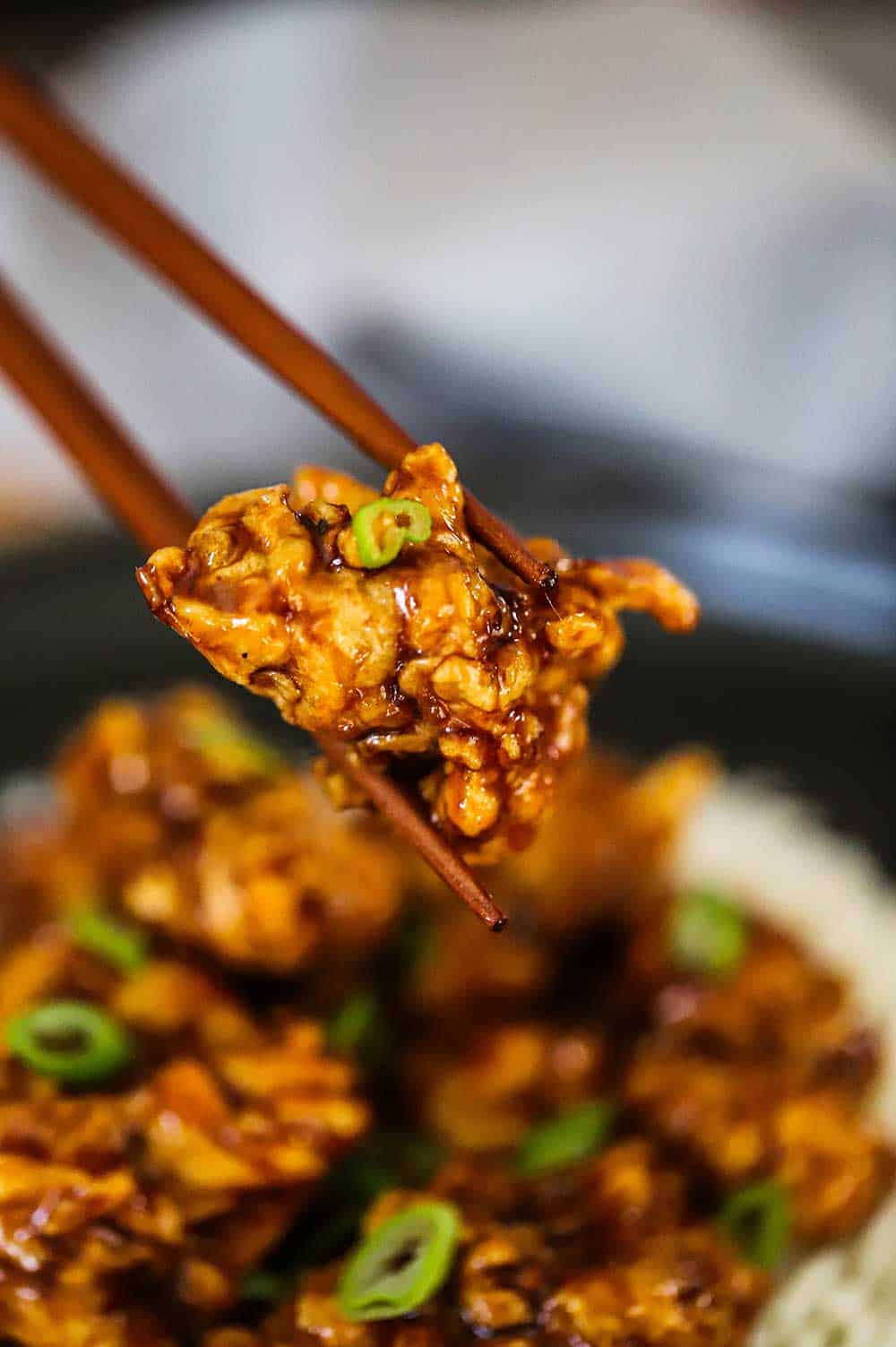 A pair of chopsticks holding up a piece of fried orange chicken over a plate of the same.