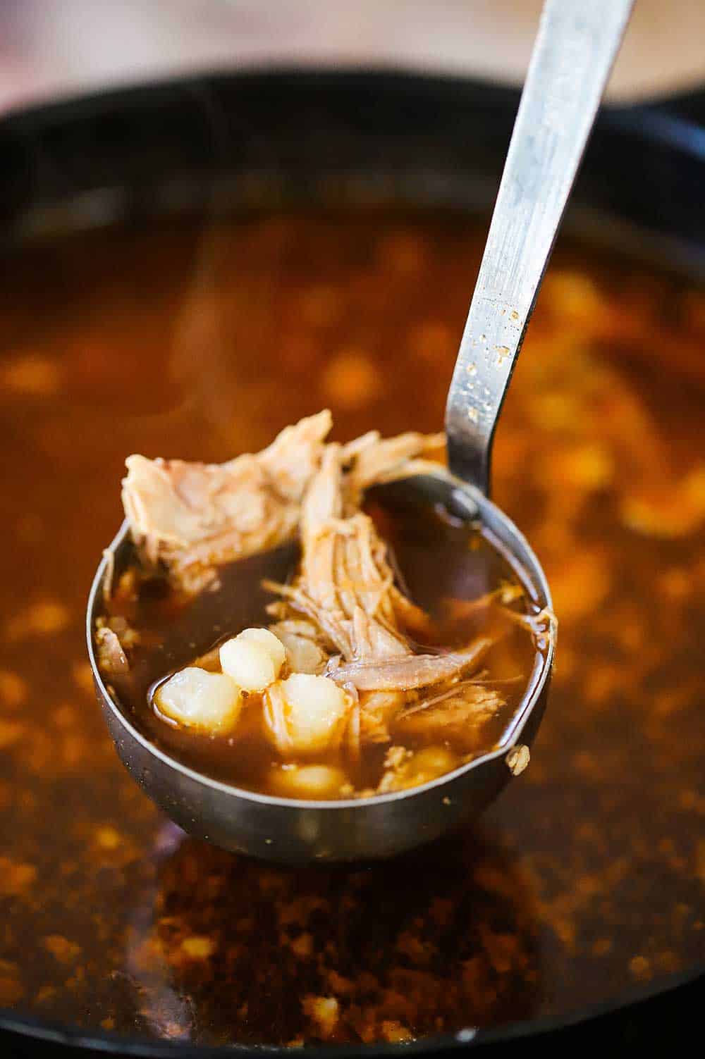 A large silver ladle filled with pork and hominy pozole over a large pot filled with the stew.