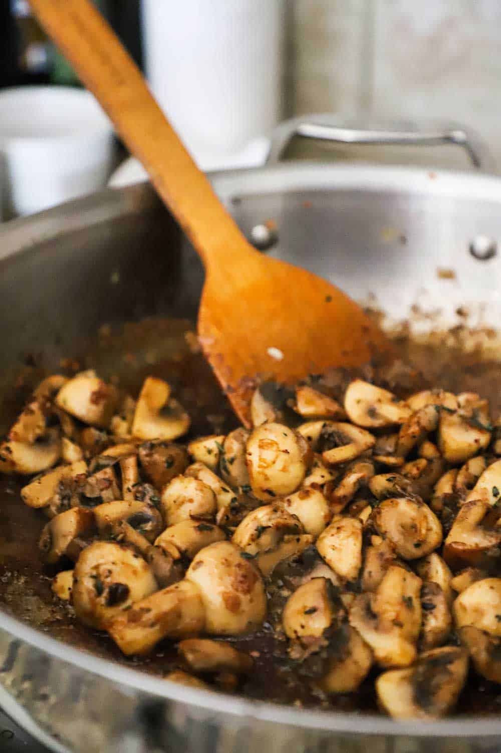 A stainless steel skillet filled with quartered wild mushrooms that have been sautéd and stirred with a wooden spoon.