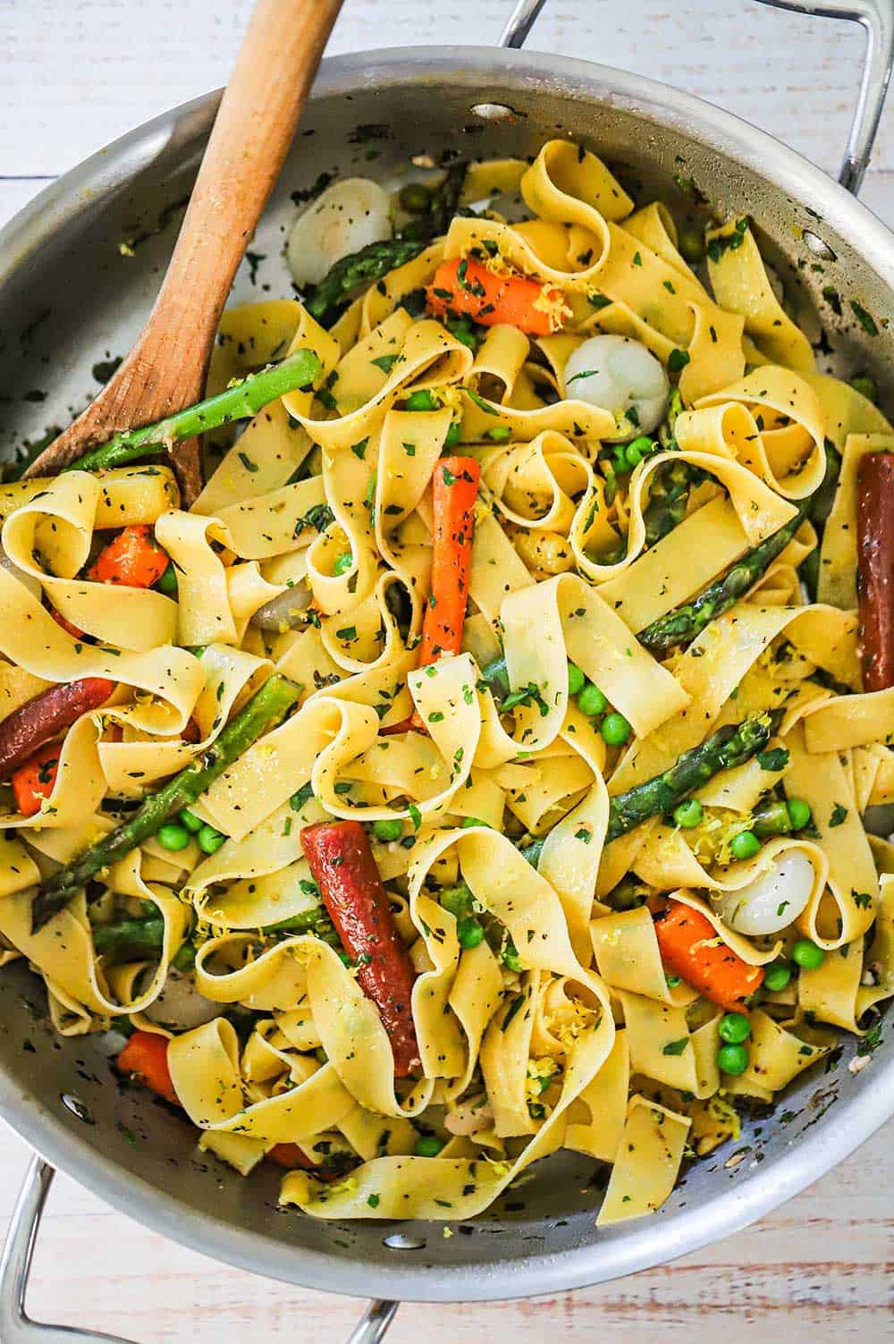 An overhead view of a large stainless steel skillet filled with pappardelle with seasonal vegetables with a wooden spoon inserted into it.