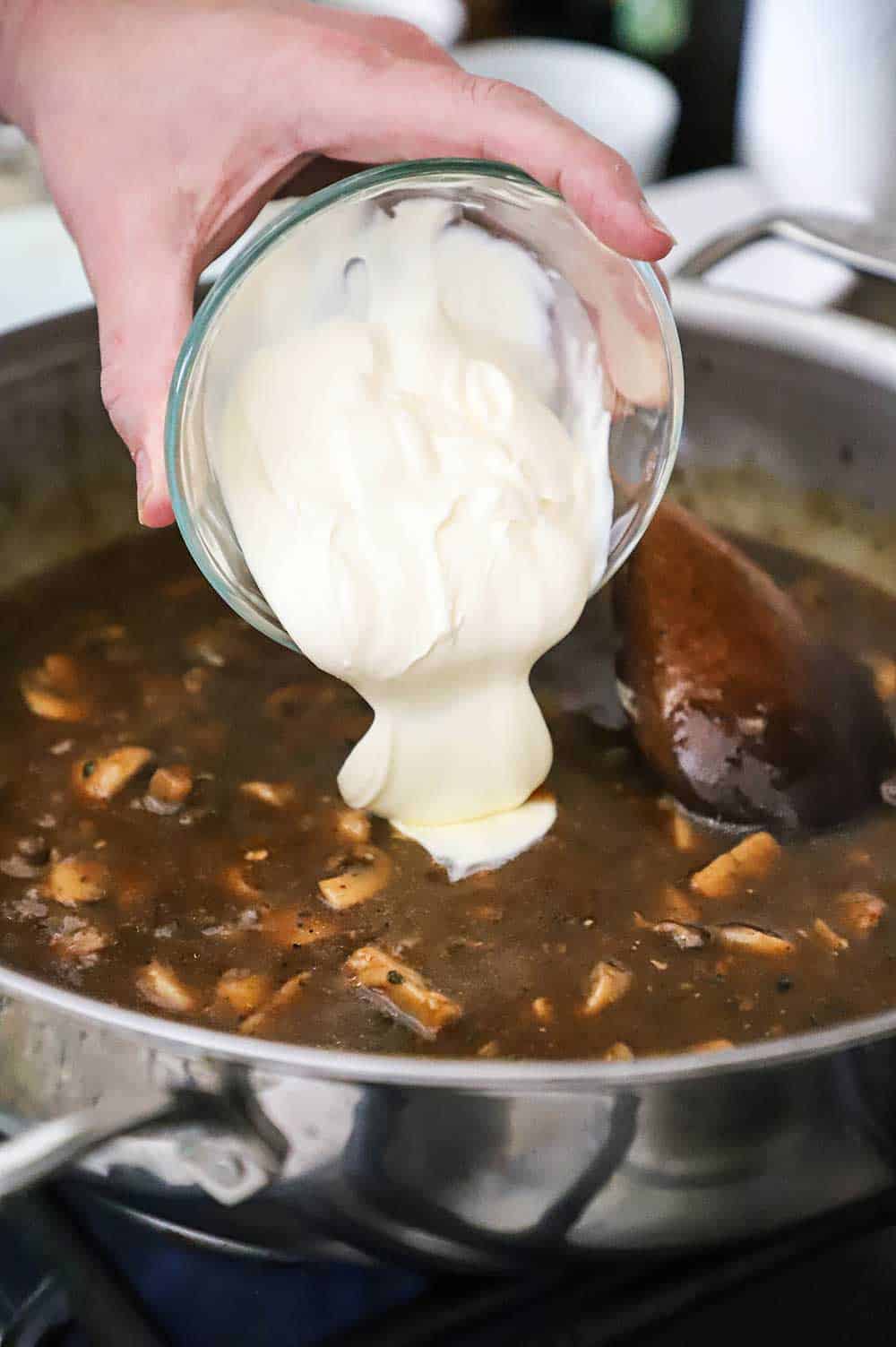 A person transferring softened crème fraîche from a glass bowl into a skillet filled with a dark brown gravy with mushrooms.