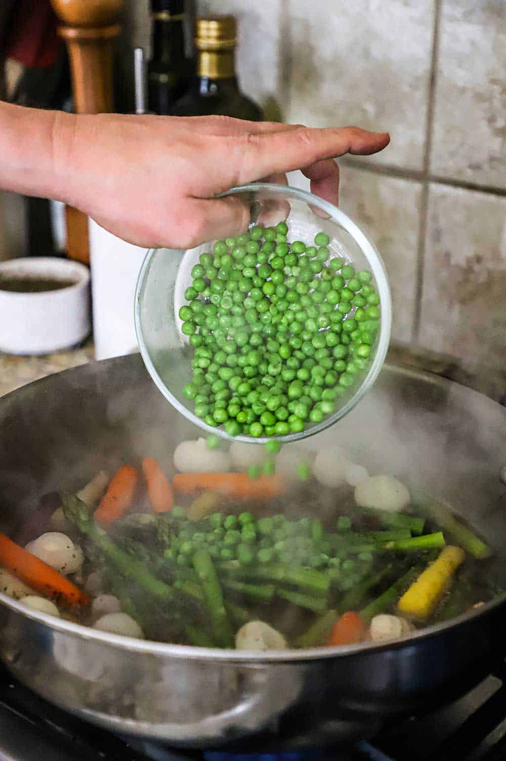 A person transferring sweet peas from a small glass bowl into a pot of simmering vegetables including carrots, asparagus, and pearl onions.
