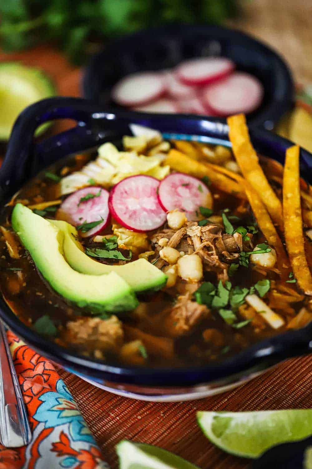A color Mexican-style bowl filled with pozole and topped with avocado slices, radish slivers, and strips of fried corn tortillas.