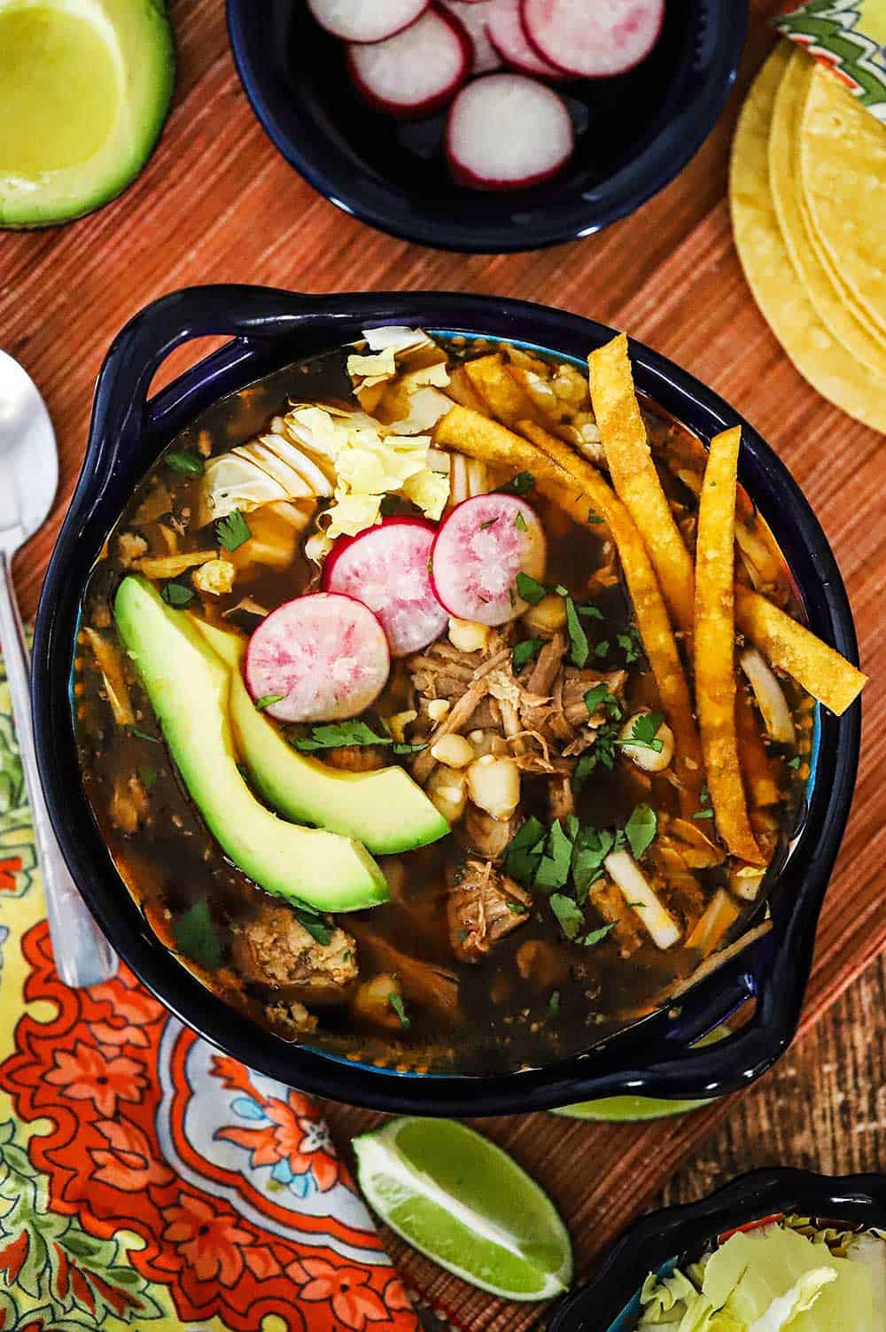 A colorful Mexican bowl filled with Mexican pozole stew surrounded by bowls of sliced radishes, cabbage, avocados, and corn tortillas.