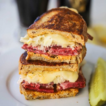A grilled rueben sandwich that has been cut in half and then stacked on top of each other on a white plate with a pickle spear nearby.