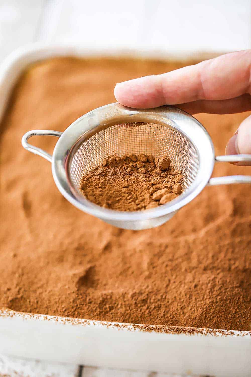A hand tapping the side of a small sieve filled with chocolate powder over a casserole dish filled with tiramisu.