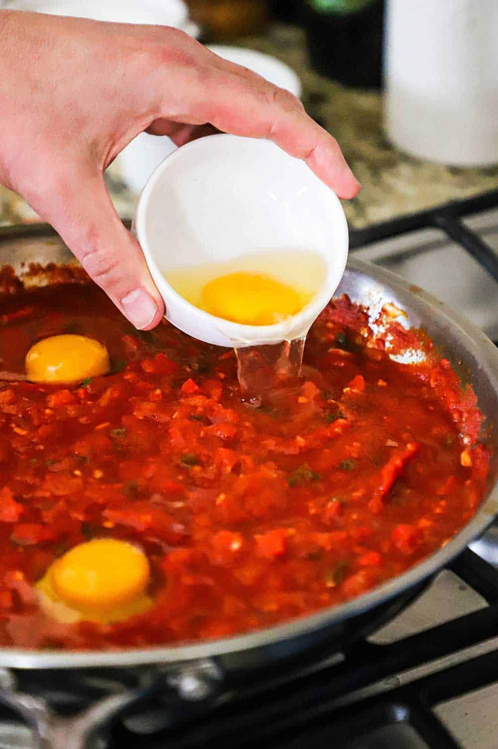 A person transferring an uncooked egg into a skillet filled with a savory tomato sauce.