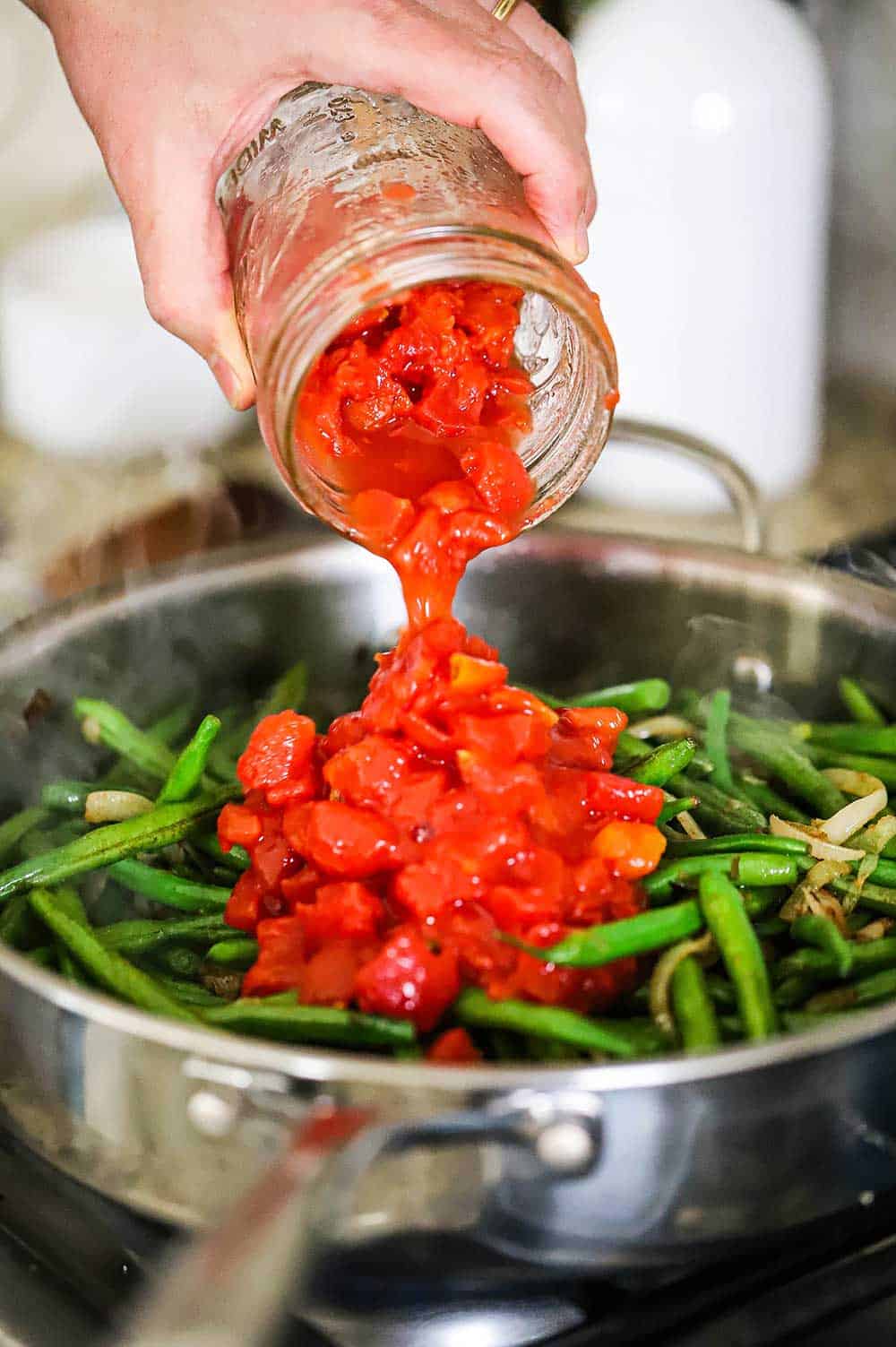A person transferring diced tomatoes into a stainless steel skillet filled with sautéed green beans and onions.