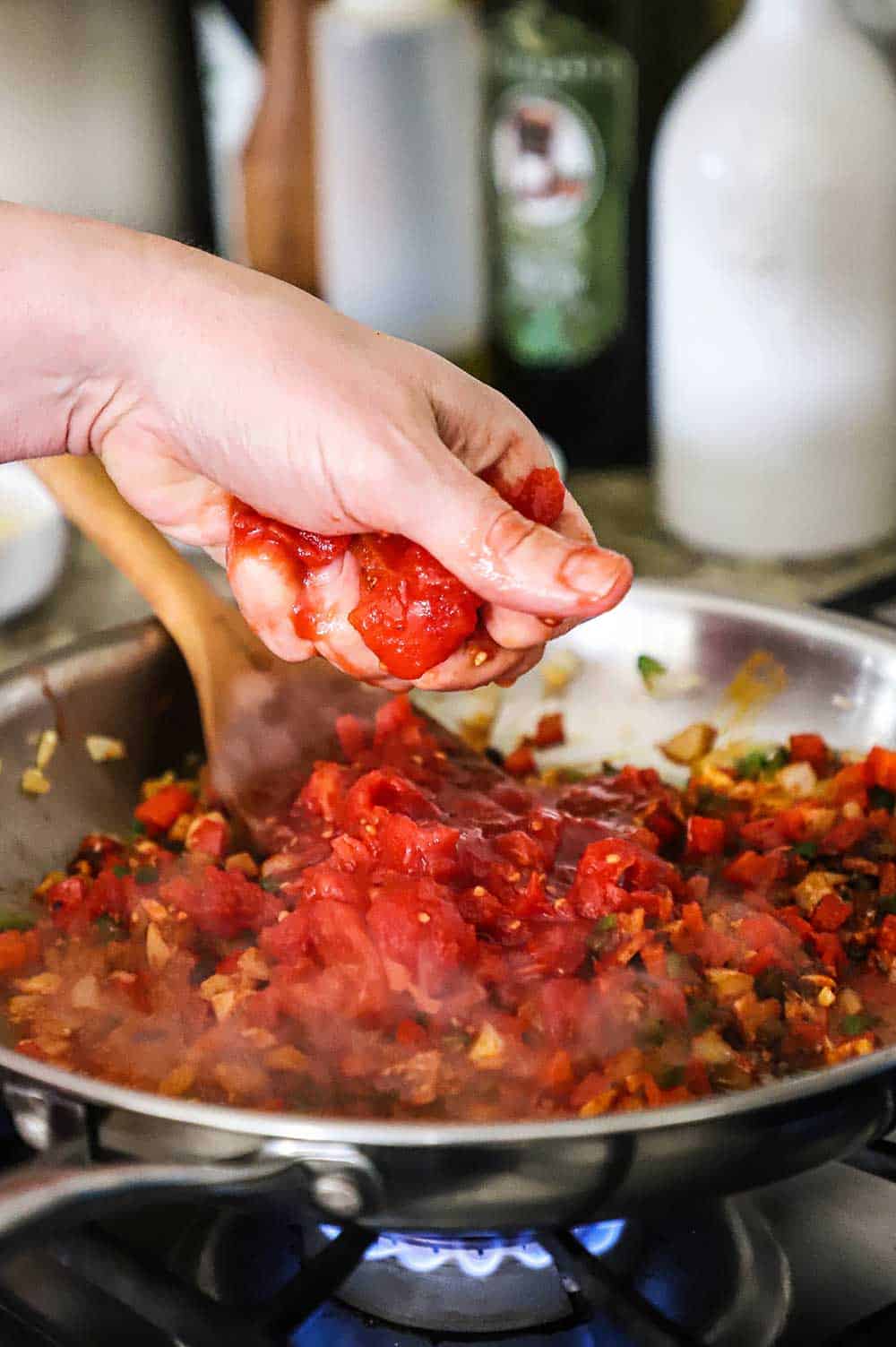 A person squeezing a whole canned tomato into a skillet filled with sautéed peppers and onions.