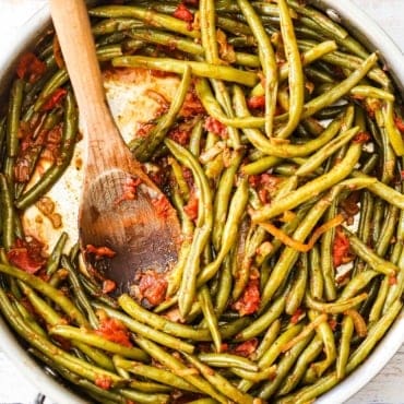 An overhead view of a stainless skillet filled with braised green beans with tomatoes with a large wooden spoon inserted into the middle.