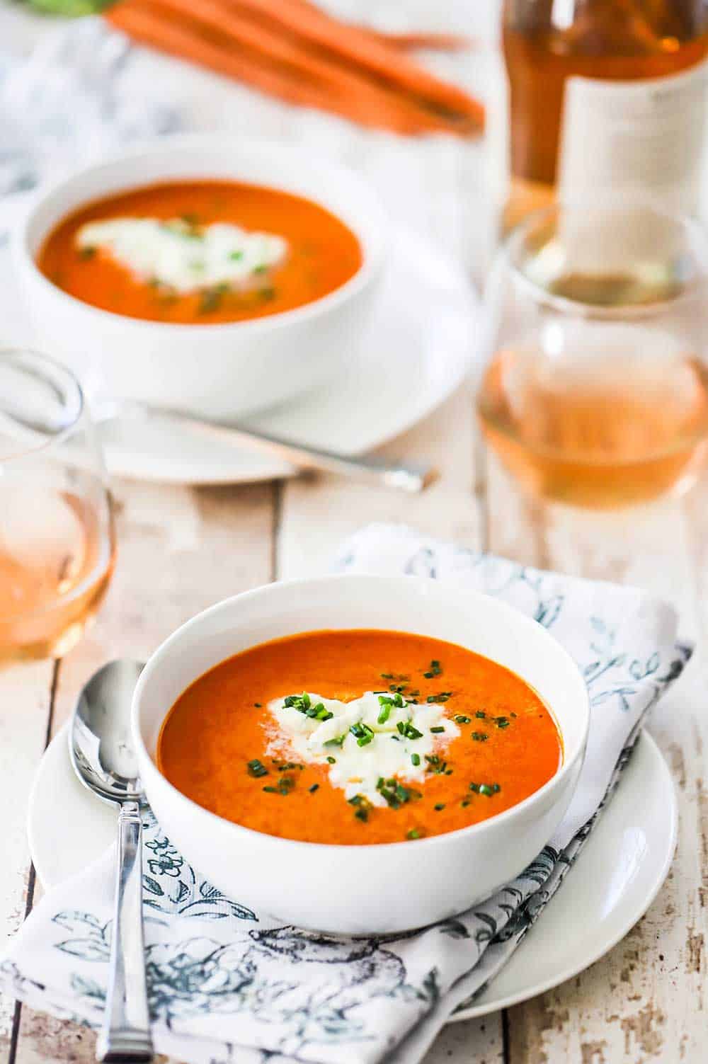 Two white soup bowls filled with carrot ginger soup sitting next to two glasses or rosé wine.