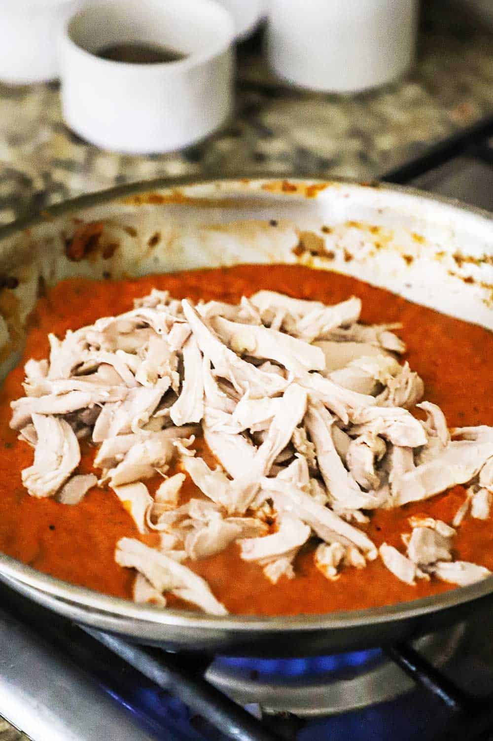 A large stainless steel skillet filled with puréed tinga sauce with shredded chicken sitting the middle of the pan. 