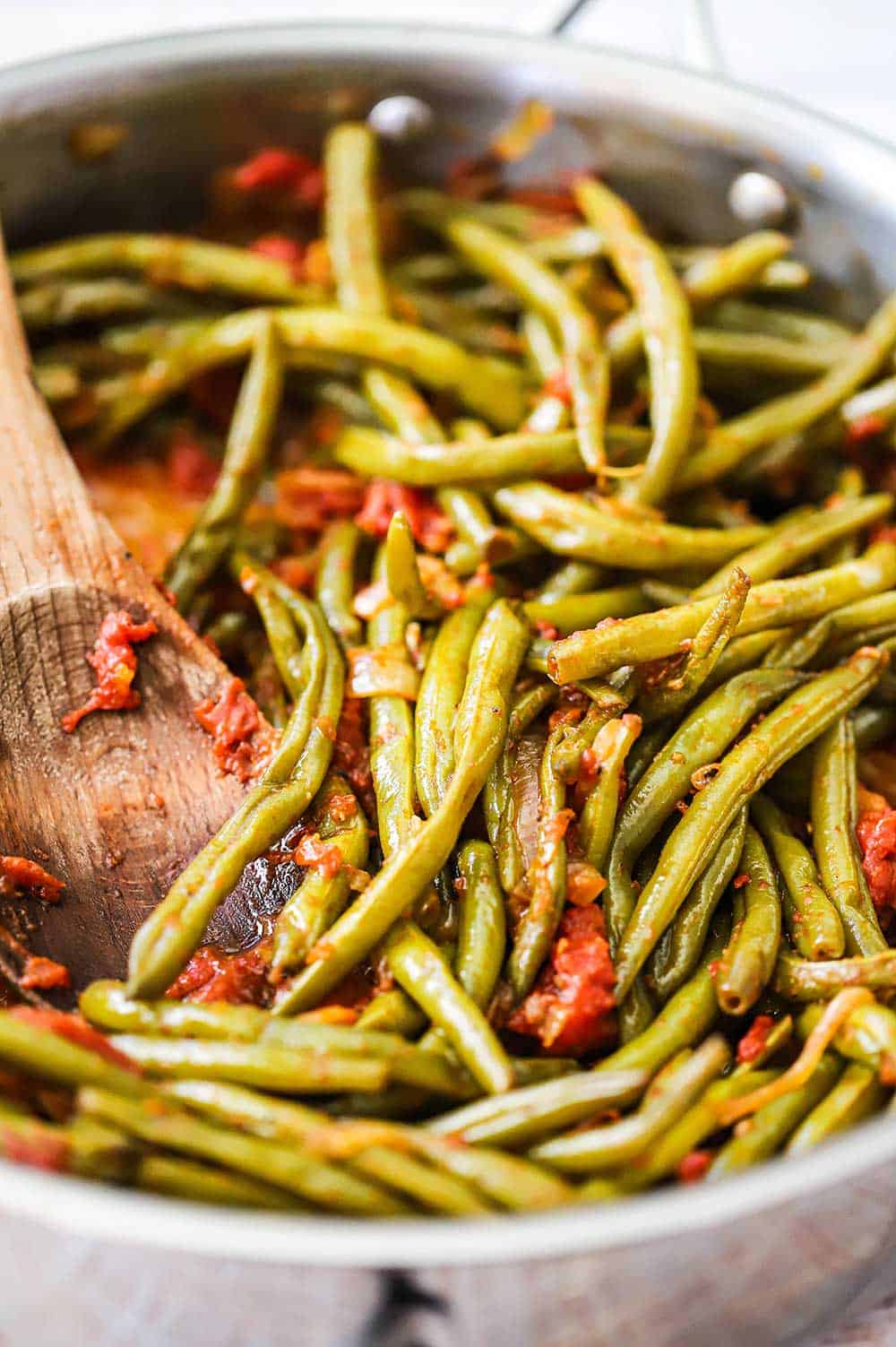 A close-up view of a wooden spoon inserted into a skillet filled with braised green beans with tomatoes.