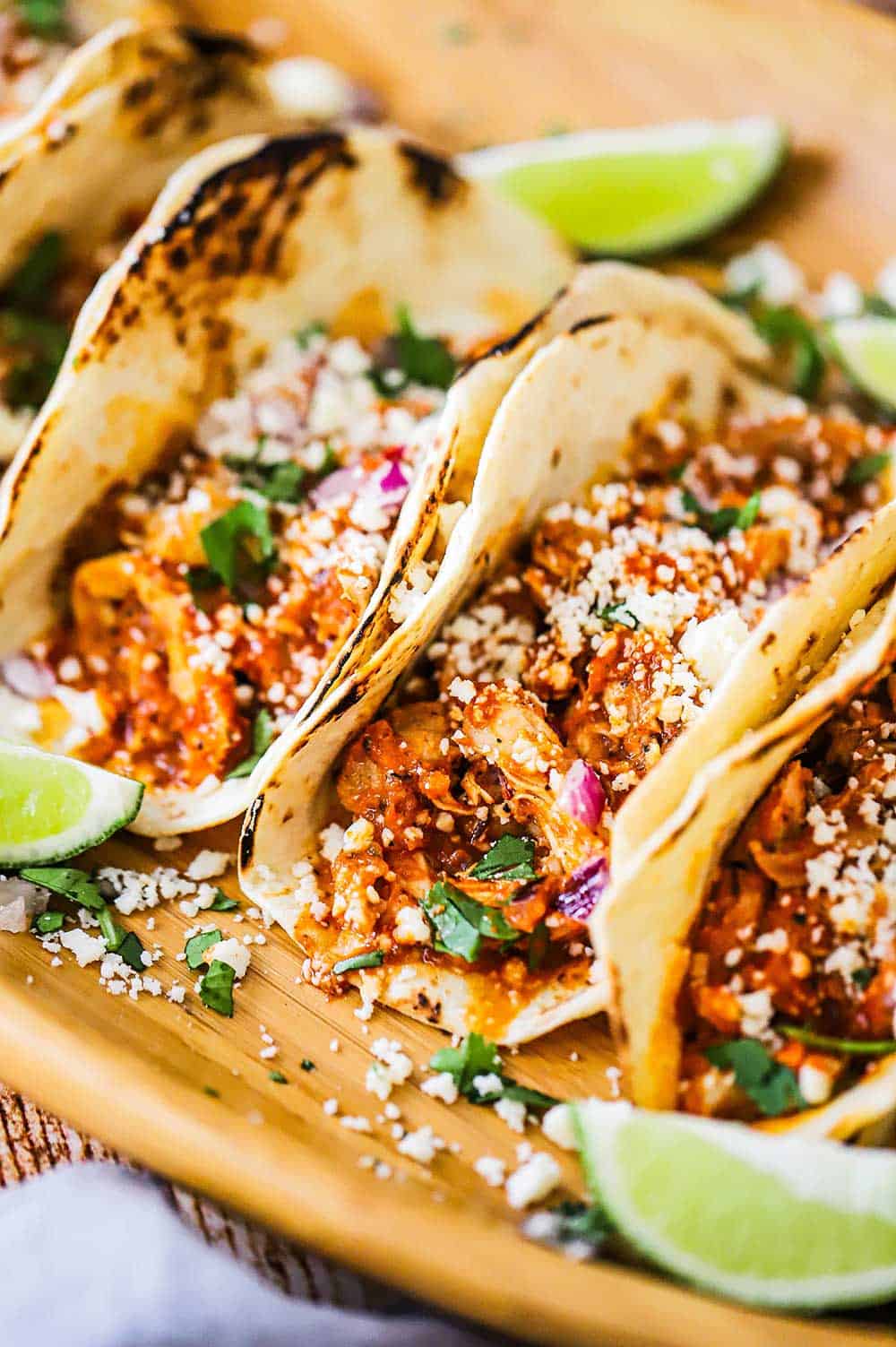 A row of easy chicken tinga tacos sitting next to each other on a wooden platter topped with crumbled cheese and garnish with lime wedges. 