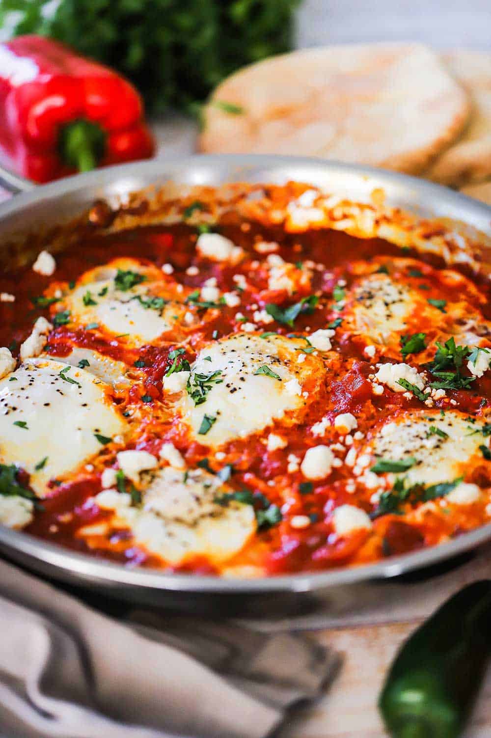 A large stainless steel skillet filled with shakshuka and a red bell pepper and pita bread sitting nearby.