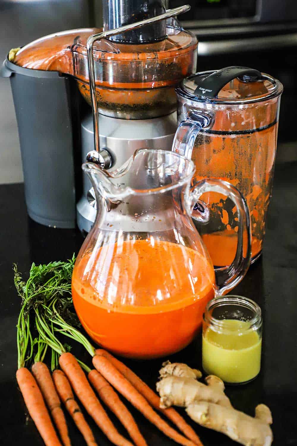 A large juicing machine that has carrot pulp in the catcher with a glass pitcher of fresh carrot juice in front of it.