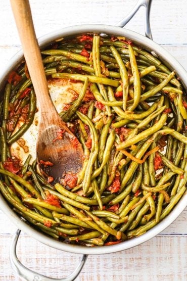 An overhead view of a stainless skillet filled with braised green beans with tomatoes with a large wooden spoon inserted into the middle.