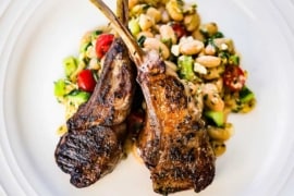 An overhead view of two marinated grilled lamb chops crisscrossing each other on a bed of a white bean salad all on a white dinner plate.