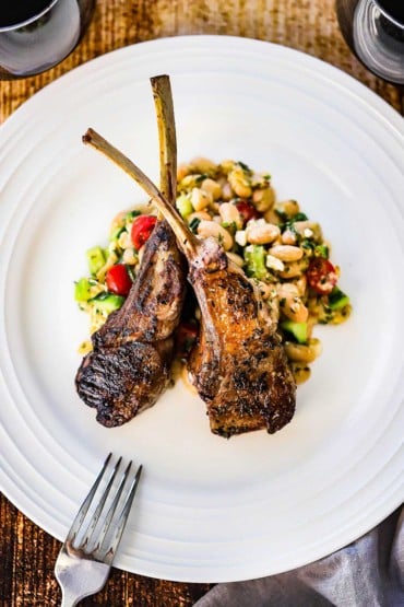 An overhead view of two marinated grilled lamb chops crisscrossing each other on a bed of a white bean salad all on a white dinner plate.