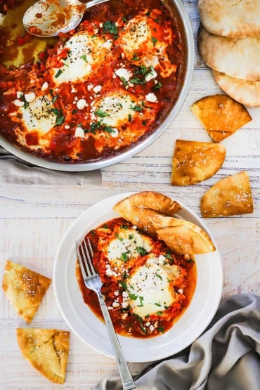 An overhead view of a large skillet filled with shakshuka with a spoon in it sitting next to a white bowl filled with poached eggs in a tomato sauce with pita triangles scattered around.