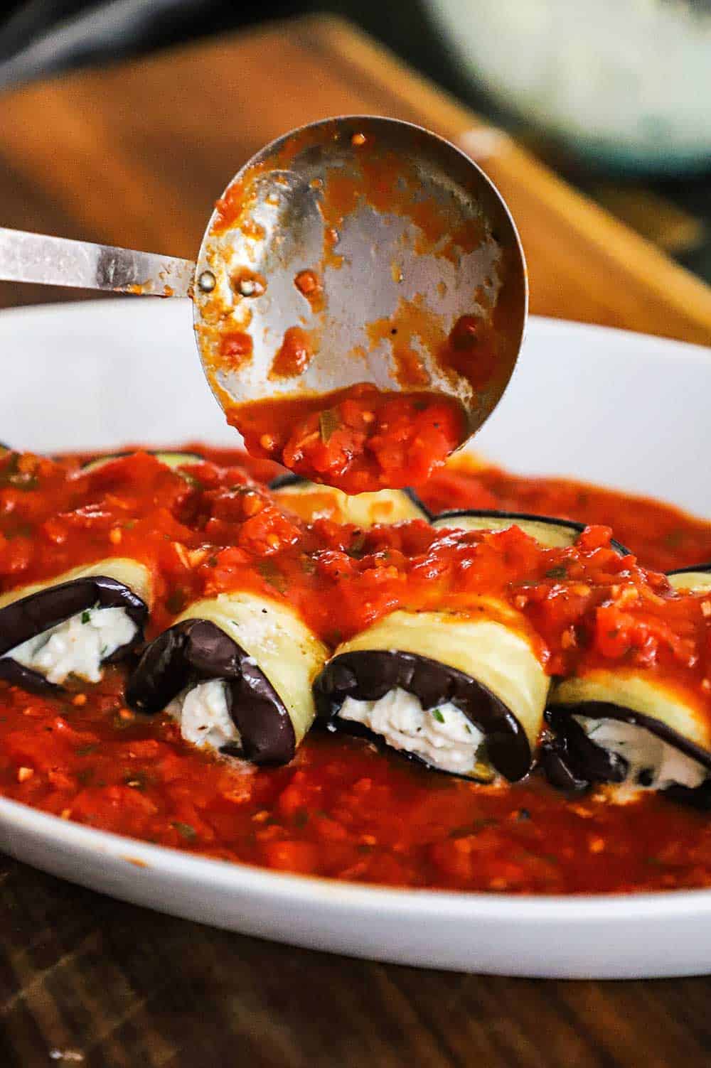 A small ladle being used to spread homemade marinara over the top of roasted eggplant that is rolled with a cheese mixture in the middle of each one.