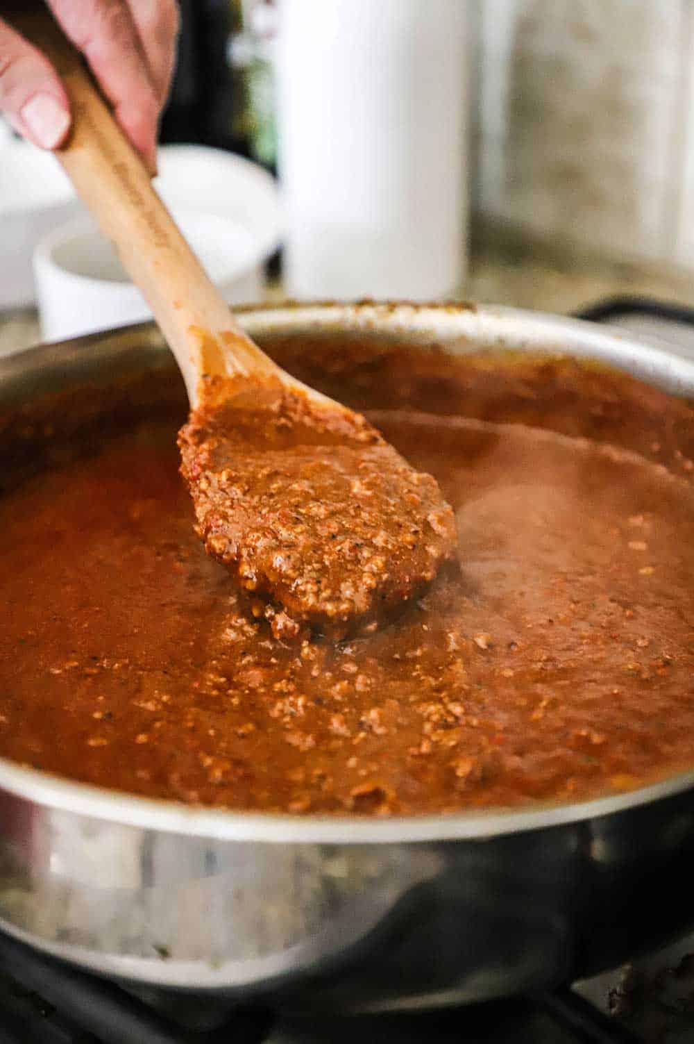 A person lifting a large wooden spoon out of a stainless steel skillet filled with homemade chili con carne sauce.