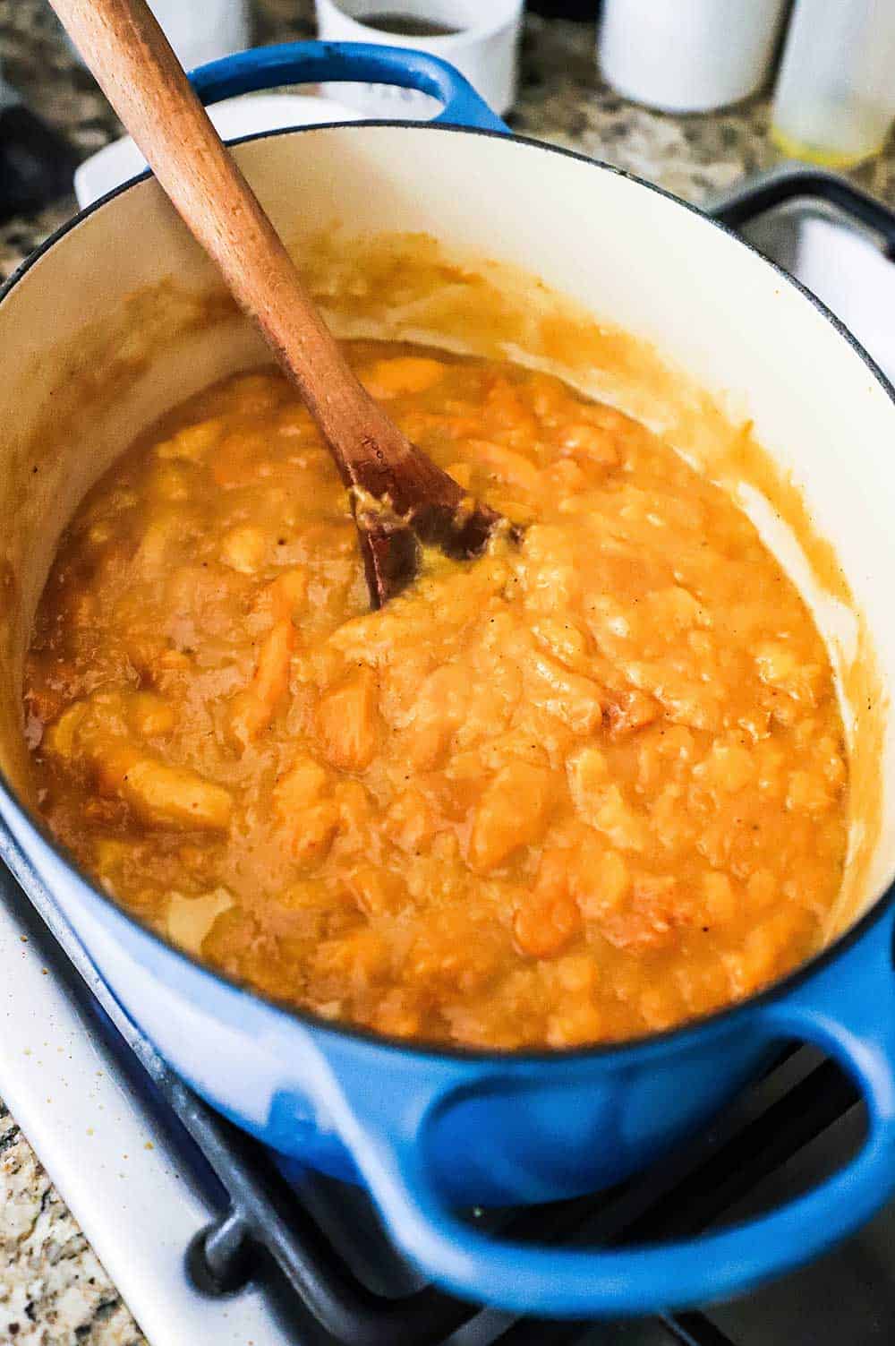 A large blue oval Dutch oven filled with peach cobbler filling and a wooden spoon stuck in the peaches.