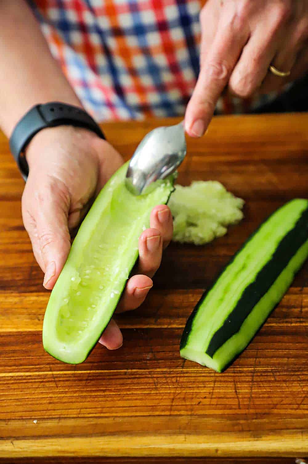 A person using a spoon to scoop out the seeds of a cucumber that has been cut in half lengthwise.