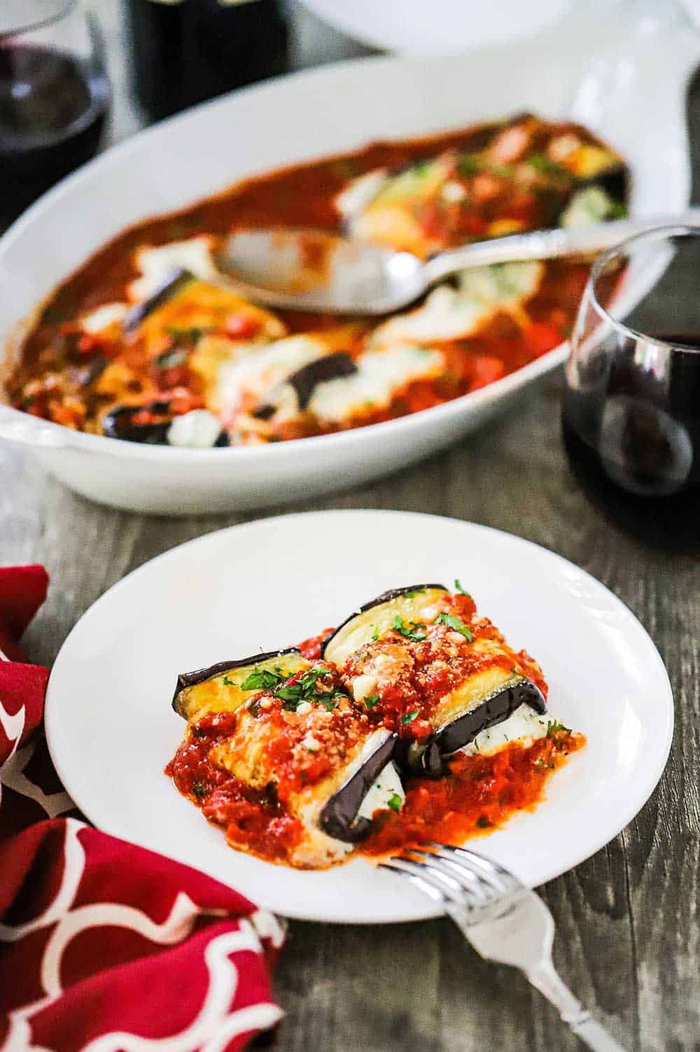 A white plate filled with two baked eggplant rollatini's sitting in front a baking dish of the same.