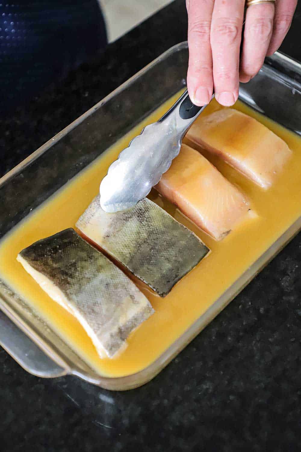A person using tongs to flip black cod fillets in a dish filled with miso mariande.