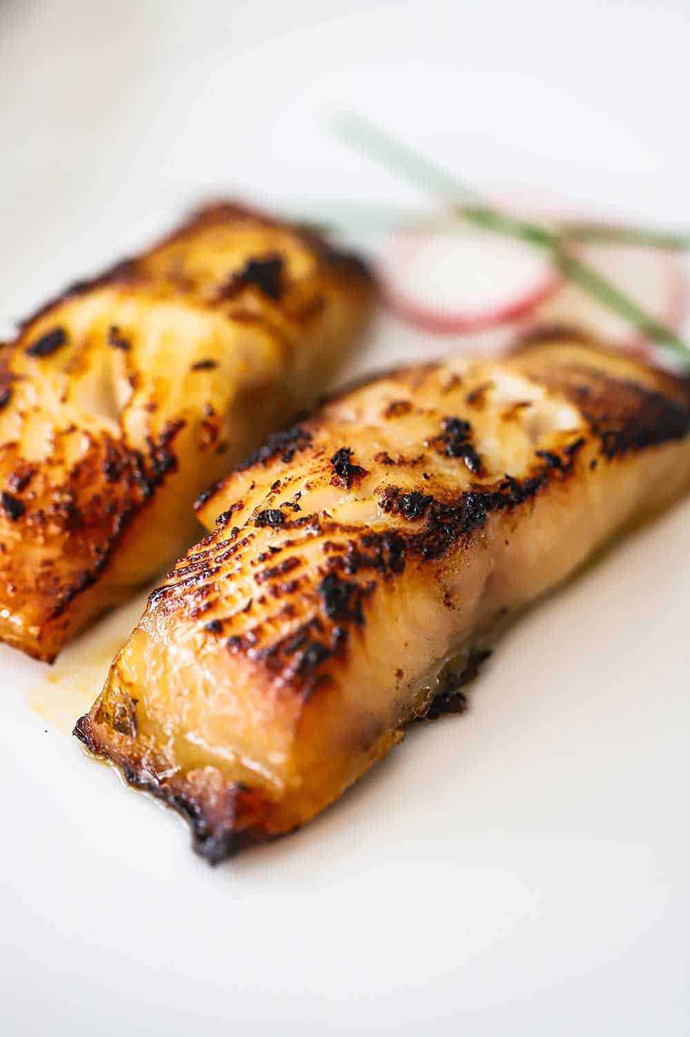 A close-up view of two miso cod fillets that have been lightly charred on a white plate.