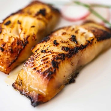 A close-up view of two miso cod fillets that have been lightly charred on a white plate.