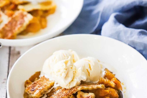 Two white bowls filled with peach cobbler and topped with scoops of vanilla ice cream with a pint of ice cream in the background.