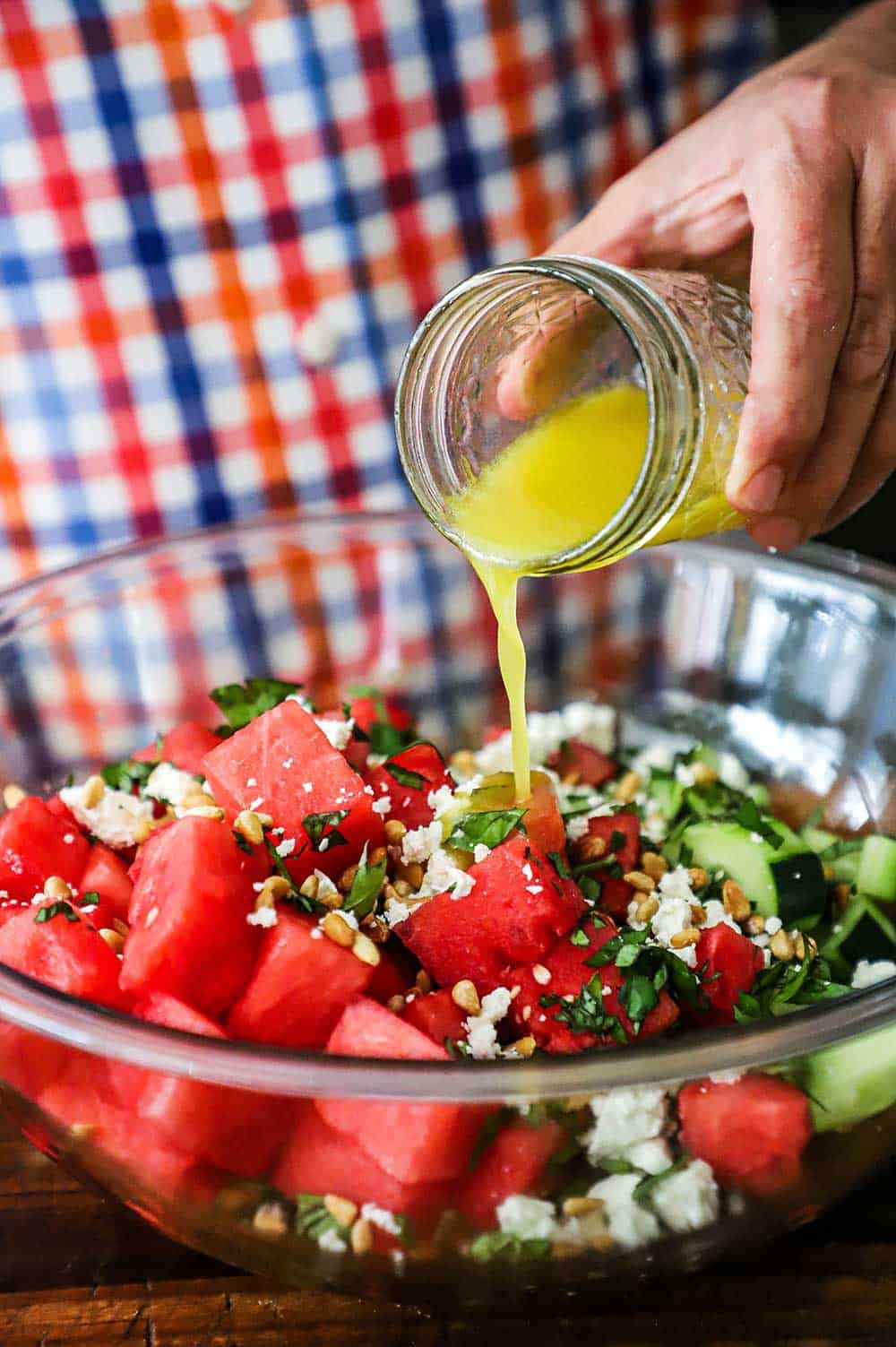 A person pouring a lemon vinaigrette into a glass bowl filled with watermelon, cucumber, feta cheese, basil, and pine nuts.