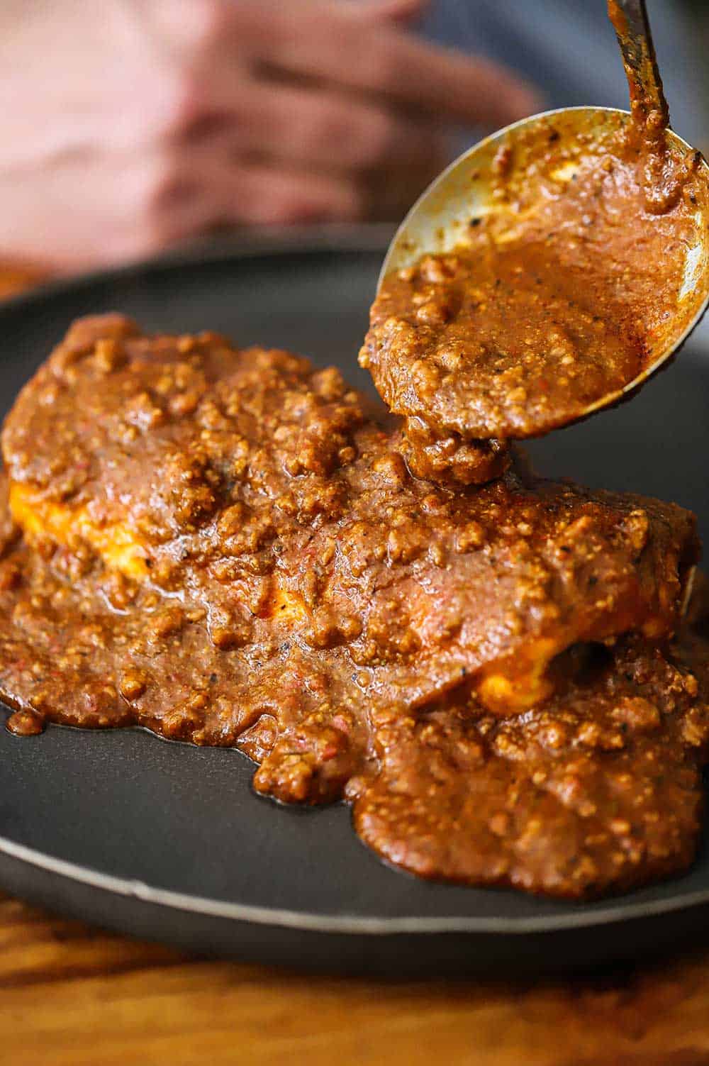 A person using a ladle to pour chili con carne sauce all over a burrito on a blue dinner plate.