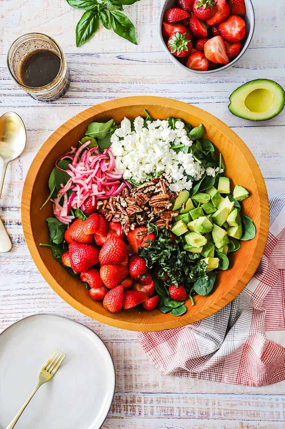 An overhead view of a large wooden salad bowl filled with halved strawberries, red onion, pecan, feta cheese, avocado chunks, and fresh spinach.