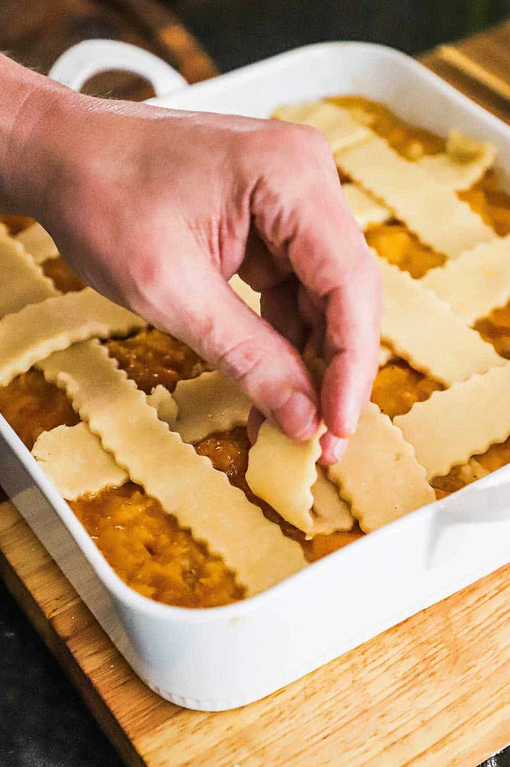 A person's hand layering cut pieces of pie dough in a lattice formation on top of a dish of old fashioned peach cobbler.