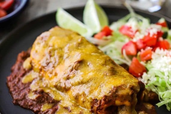 A blue plate filled with a smothered beef burrito with chili con carne sitting next to a salad topped with diced tomatoes.