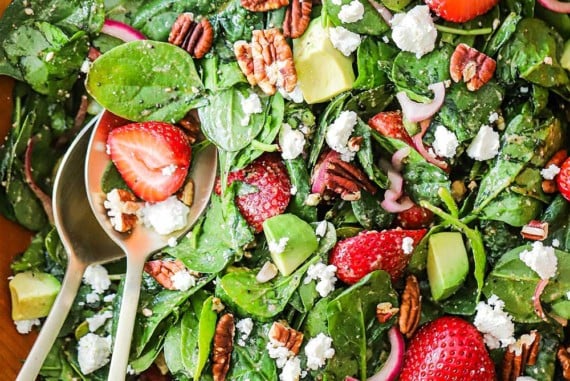 An overhead view of a fresh strawberry spinach salad with avocado that has been tossed together in a wooden bowl.